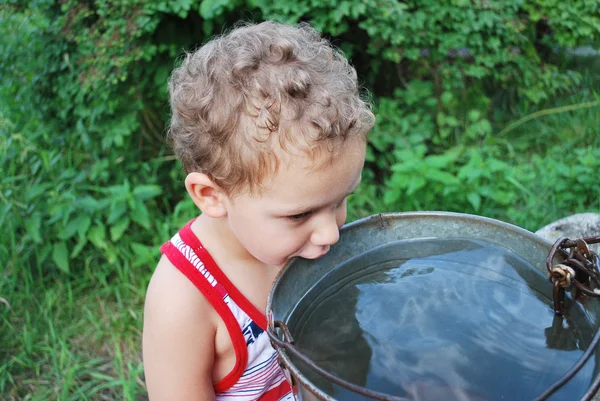 Im Dorf in der Nähe des Brunnens trinkt ein kleiner Junge Wasser aus einem — Stockfoto