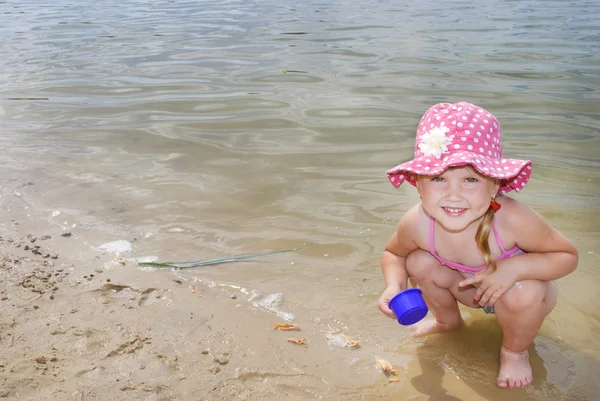Na pláži nedaleko jezera v písku holčička hraje s — Stock fotografie