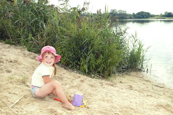 The beach at the lake in the sand a little girl in a hat playin — Stock Photo, Image