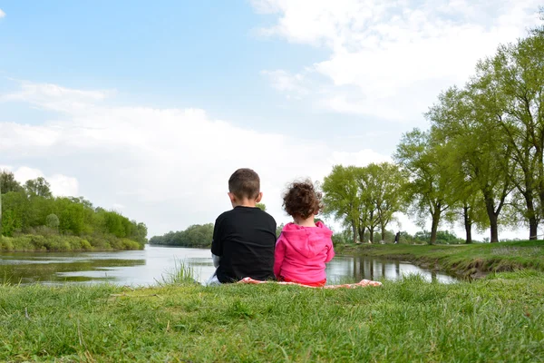 Irmão e irmãzinha sentados na margem do rio . — Fotografia de Stock