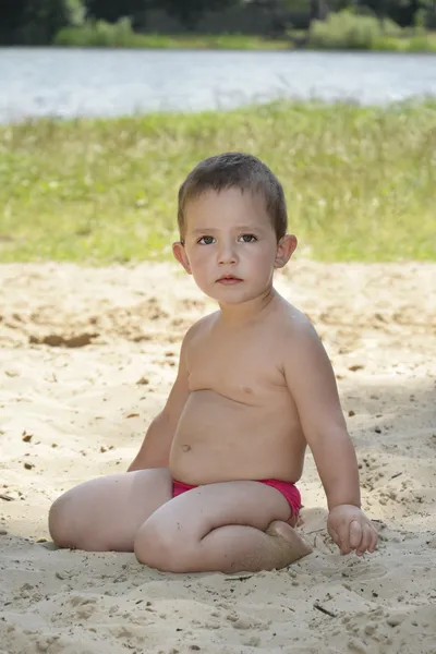 Klein meisje zit op een strand in het zand in de buurt van het meer. Stockafbeelding