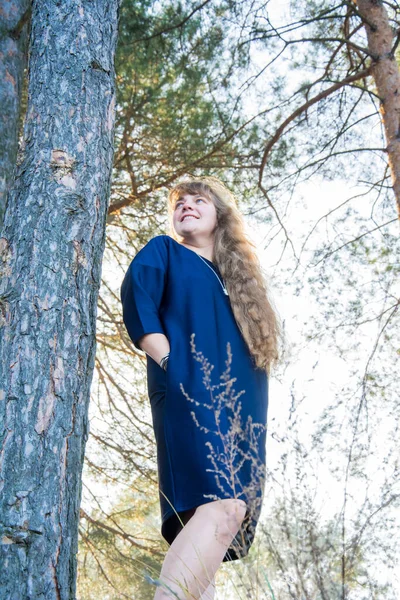 秋の午後には 長い波状の髪と青いドレスを着た美しい女の子が松の木の近くに立っています — ストック写真