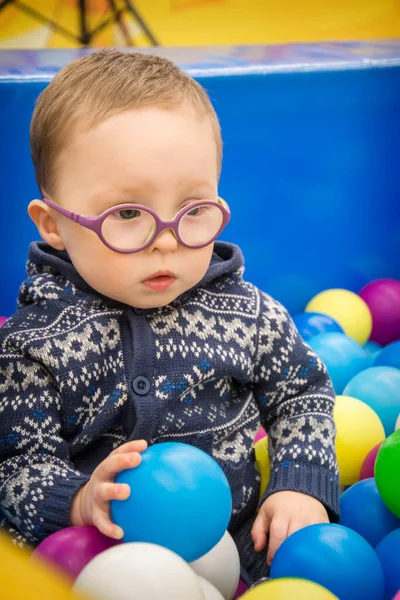 青いセーターに眼鏡をかけた小さな男の子がプレイルームで多色のプラスチックボールで遊んでいます 手に持っている — ストック写真