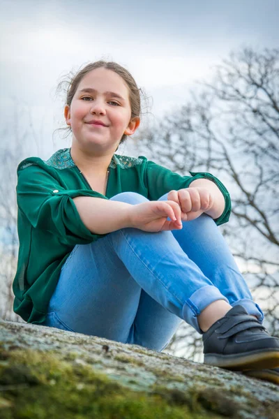 On a bright day in the spring, a girl with a scythe sits on the edge of a cliff in blue jeans and a green shirt, side view.