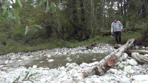 Een man in een geruite jas steekt de rivier over en balanceert op een boomstam. — Stockvideo