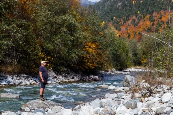 Sonbaharda bir dağ nehrinin yanındaki bir kayanın üzerinde duran şapkalı bir adam dağların yamaçlarına bakar.