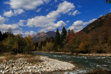 Bir dağ zirvesi, berrak bir dağ nehri ve güzel güneşli bir günde mavi gökyüzünde beyaz bulutlar. Laba Nehri, Zakan Tepesi. Ekim