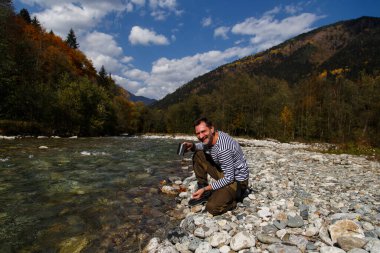 Adam nehirden su içiyor. Bir adam temiz bir dağ nehrinden su içer. Metal bir turist bardağını suyla doldurdu.