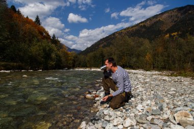 Adam nehirden su içiyor. Bir adam temiz bir dağ nehrinden su içer. Metal bir turist bardağını suyla doldurdu.