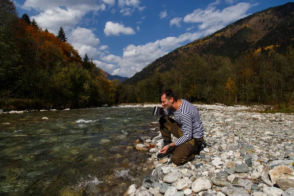 Adam nehirden su içiyor. Bir adam temiz bir dağ nehrinden su içer. Metal bir turist bardağını suyla doldurdu.