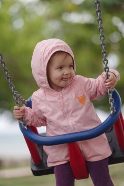 2 yaşında beyaz bir kız çocuk parkında oynuyor, salıncakta sallanırken eğleniyor.