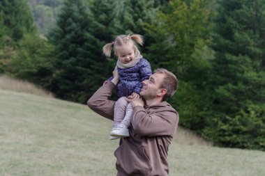 Father lifting a cheerful toddler daughter outdoors in nature clipart