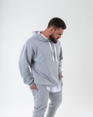 Gri spor kıyafetli sakallı erkek model stüdyoda beyaz arka plana bakıyor.