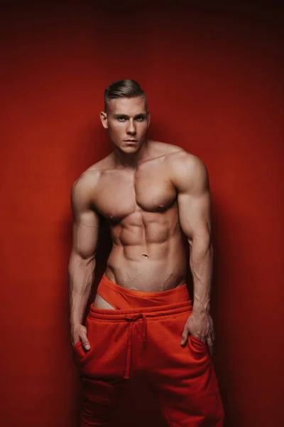 Sexy blonde in red sweatpants posing in studio. Muscular young man in sportswear on red background. Handsome guy with six pack abs.