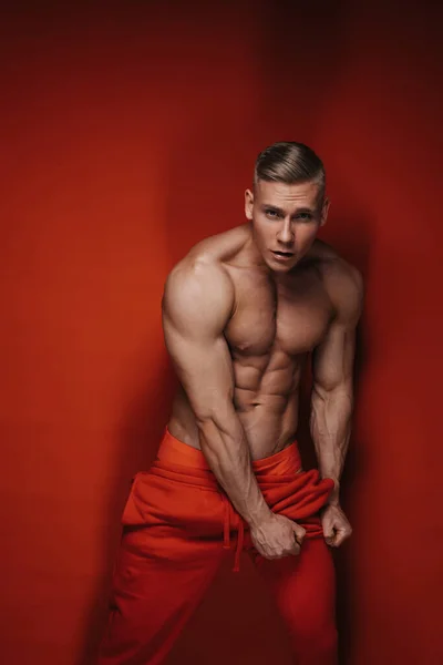 Sexy blonde in red sweatpants posing in studio. Muscular young man in sportswear on red background. A handsome six pack abs guy is pulling up his left pant leg.