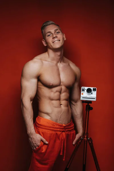 Shirtless young and handsome male model in red pants near tripod with camera at red background. Sexy photographer. Fitness model in studio.
