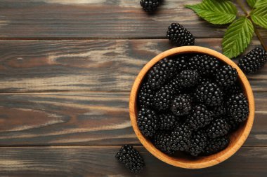 Kahverengi ahşap arka planda, ahşap bir kasede yapraklarla dolu olgun böğürtlenler. Üst görünüm.