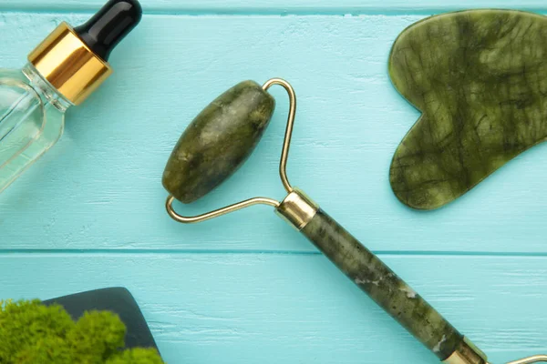 Green jade roller and gua sha stone for facial massage and on blue background. Home beauty and selfcare accessories. Face roller for anti age wrinkle treatment.