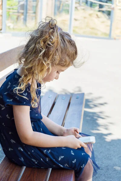 幼児の女の子はベンチの上にハンチングし 彼女の携帯電話を見て座ります 悪い体の姿勢 — ストック写真