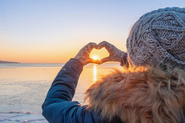 冬服の女の子は手袋をして手で心を作る 日没時の凍結した海の背景に 自然への愛 ストック画像