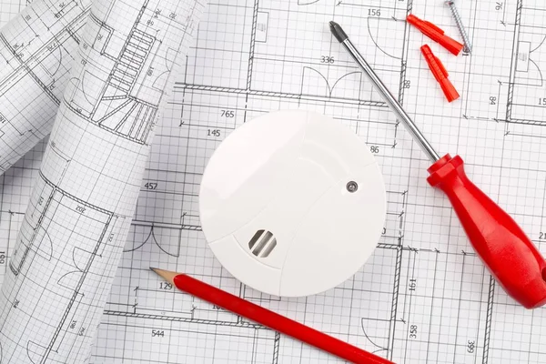 Smoke detector or fire alarm sensor with rolls of architectural drawing background with tools and screws, house safety or security concept, flat lay top view from above