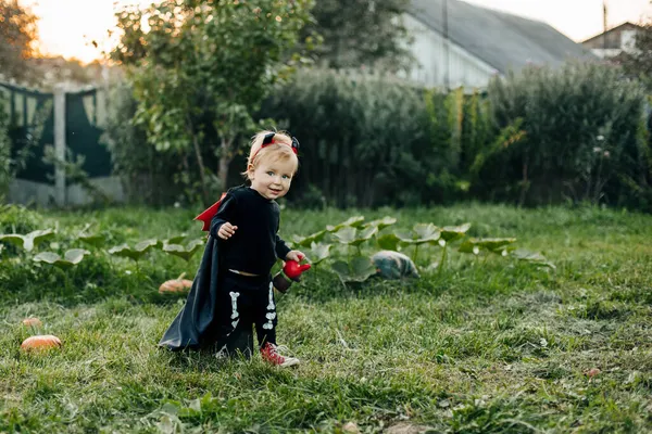 Un niño encantador con un disfraz de diablo y un impermeable se cuela alrededor del jardín. concepto de Halloween, disfraz de carnaval — Foto de Stock