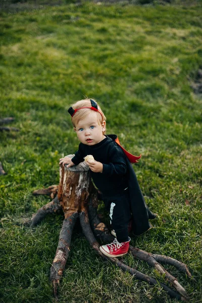 Um menino loiro com um traje de diabo com um manto vermelho fica perto de um toco de árvore. Véspera de Halloween, traje de carnaval, tradições — Fotografia de Stock