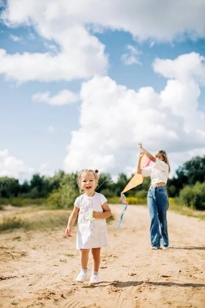 Uma garotinha rindo e sua mãe correm ao longo da estrada para o campo e voam um papagaio. Mãe e filha se divertir juntos — Fotografia de Stock