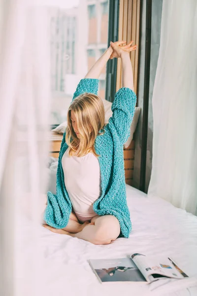 Uma jovem de pijama e um casaco azul de malha está sentada de pernas cruzadas na cama, esticada. A menina gosta da manhã e desfruta do novo dia — Fotografia de Stock