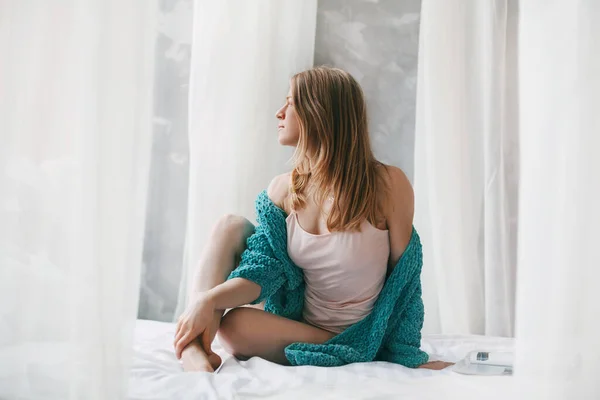 Aranyos lány, aki keresztbe tett lábbal ül az ágyon reggel. Fiatal nő pizsamában ül az ágyon, miután felébredt. — Stock Fotó