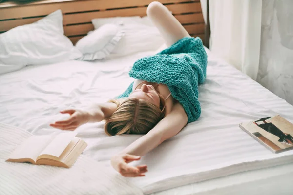 Uma linda menina deita-se na cama no início da manhã e se estende depois de acordar, ao lado de um livro aberto — Fotografia de Stock