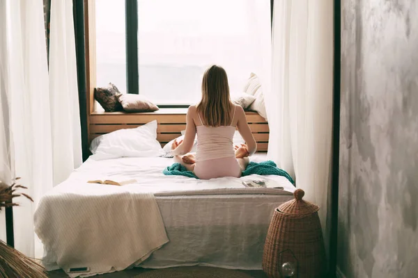 Uma jovem mulher de pijama está sentada em uma cama em uma posição de lótus com os dedos dobrados em um gesto de mudra, desfrutando de uma manhã de meditação profunda mindfulness. Vista traseira — Fotografia de Stock