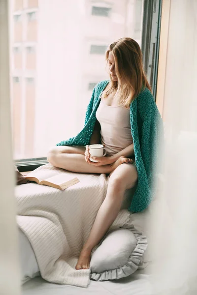 한 소녀가 이른 아침에 창가에 앉아 뜨거운 커피를 마시고 있는데, 책이 열려 있는 책이 옆에 놓여 있습니다 — 스톡 사진