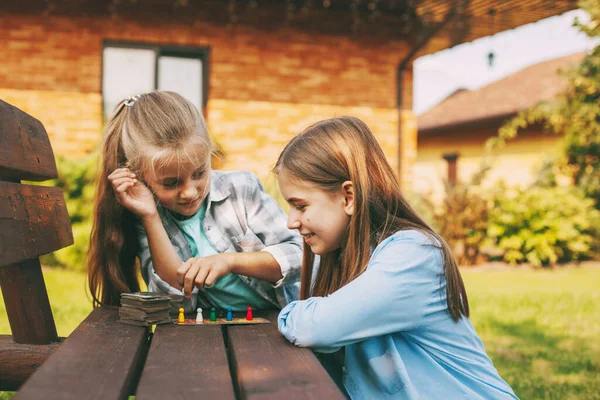 Dos hermanas se ríen y juegan un juego de mesa de madera con coloridas fichas azules, rojas, verdes y amarillas al aire libre fuera de su casa. —  Fotos de Stock