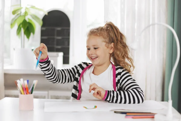 Una chica alegre toma una pluma de fieltro de color de un vaso y dibuja un arco iris en un album.Home juegos, tareas, entrenamiento en casa, la distancia social —  Fotos de Stock