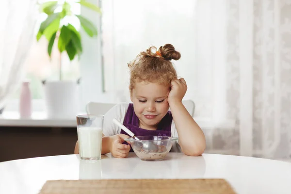Carina bambina mangia cereali con latte per colazione. La ragazza guarda attentamente la ciotola con. Colazione sana, prendersi cura dei bambini. Spazio per testo — Foto Stock