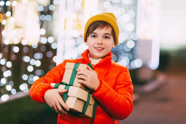 Un niño feliz en una chaqueta naranja con una caja de regalo en sus manos en un mercado de Navidad en una noche de invierno. Regalos de Navidad, preparación para las fiestas — Foto de Stock