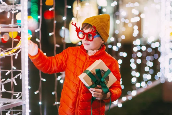Un niño alegre con una chaqueta naranja y gafas divertidas camina por la ciudad de la noche con iluminación festiva. Regalos de Navidad, preparación para las fiestas — Foto de Stock