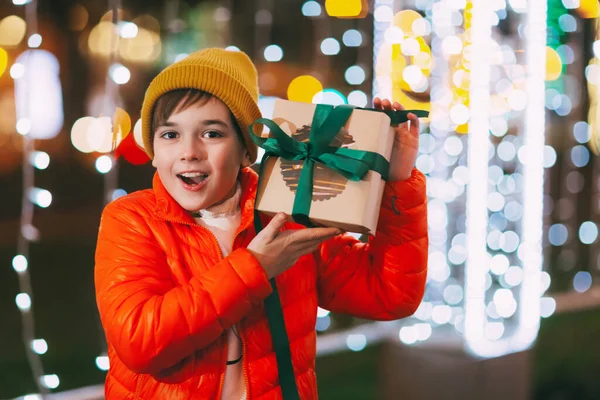 Un niño feliz en una chaqueta naranja con una caja de regalo en sus manos en un mercado de Navidad en una noche de invierno. Regalos de Navidad, preparación para las fiestas —  Fotos de Stock