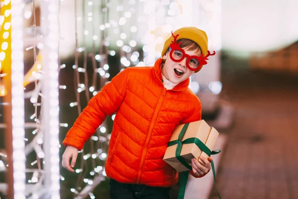 Un niño alegre con una chaqueta naranja y gafas divertidas camina por la ciudad de la noche con iluminación festiva. Regalos de Navidad, preparación para las fiestas — Foto de Stock