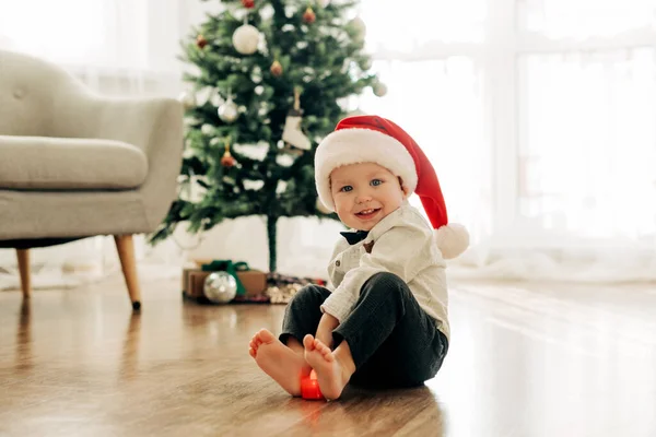 Seorang anak laki-laki tertawa dengan topi Santa Claus duduk di lantai dengan latar belakang pohon Natal Stok Lukisan  