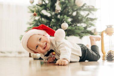 Elinde lolipopu olan bir Noel çocuğu bir Noel ağacının arka planında yere yatar ve güler. Tatil için hazırlık, çocuklar neşe, pozitif ruh hali.