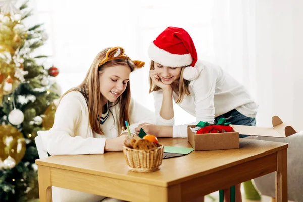 Två charmiga flickor skriver hälsningar och önskar vänner och släktingar på julafton eller nyårsafton nära julgranen — Stockfoto