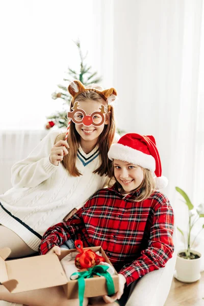 Dos hermosas adolescentes en trajes de Navidad abren regalos en casa. Humor de Navidad, regalos de Navidad. El concepto de unas vacaciones de Año Nuevo en casa —  Fotos de Stock