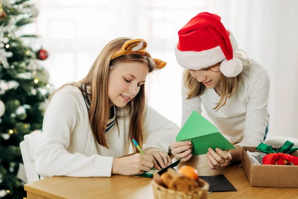 Due ragazze affascinanti scrivono saluti e auguri ad amici e parenti la vigilia di Natale o Capodanno vicino all'albero di Natale. — Foto Stock