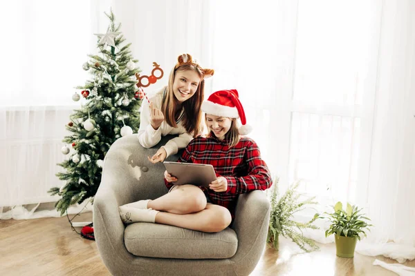 Due sorelle gioiose sono sedute a casa vicino all'albero di Natale e si congratulano con i loro amici e nonni per Natale e Capodanno e scherzano. Saluti virtuali, congratulazioni online — Foto Stock