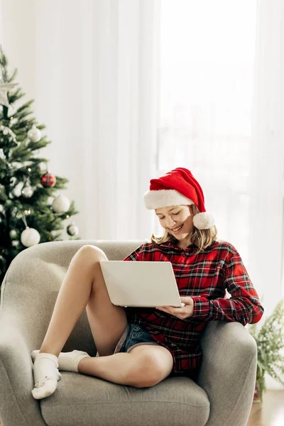 Feliz adolescente con sombrero de Santa Claus usando el ordenador portátil en la sala de Navidad decorada con interiores charlando con amigos y familiares —  Fotos de Stock