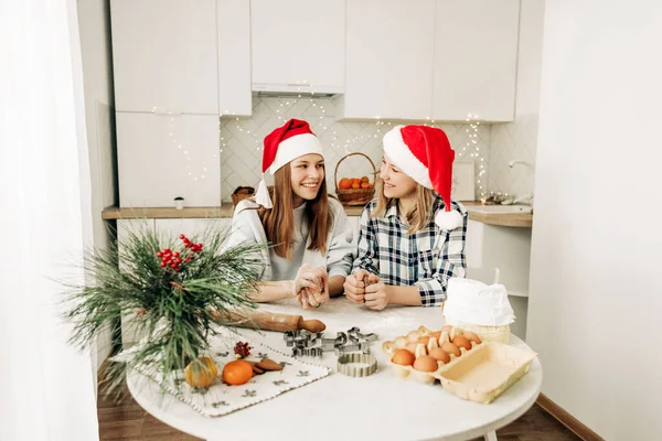 Two charming sisters in Santa Claus hats bake cookies for Christmas in the kitchen. Christmas cooking, preparation for the holiday, Christmas cookies — Stock Photo, Image
