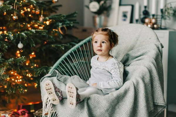 A little girl is sitting in an armchair and smiling against the background of a Christmas tree on New Years Eve. Christmas mood, expectation of magic — Stock Photo, Image