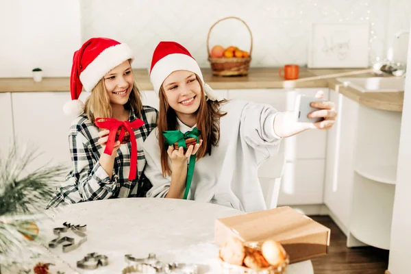 Ritratto di due sorelle carine in un cappello di Natale di Babbo Natale in cucina, scattare selfie con uno smartphone, mentre si preparano regali la vigilia di Natale e Capodanno — Foto Stock